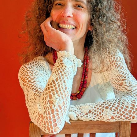 Eine Frau mit lockigem Haar trägt einen weißen gehäkelten Pullover und eine rote Perlenkette. Sie sitzt auf einem Holzstuhl mit einer roten Wand im Hintergrund.