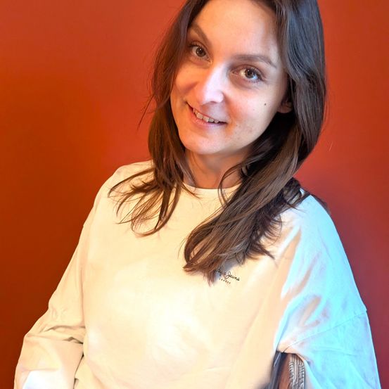 Eine Frau mit braunen Haaren, lächelnd, steht vor einer roten Wand. Sie trägt ein weißes Langarmshirt mit Text auf der linken Brustseite.