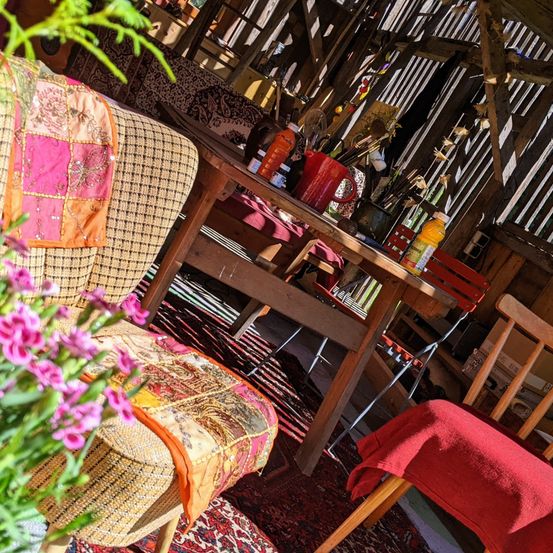 Ein gemütlicher Raum mit einem Holztisch und Stühlen. Bunte Kissen, Blumen und Flaschen stehen auf dem Tisch. Der Raum hat Holzwände und einen Teppich auf dem Boden.