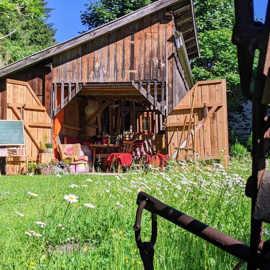 Eine rustikale Holzscheune mit offenen Türen, gefüllt mit Möbeln und Werkzeugen, umgeben von einer Wiese mit Gänseblümchen.