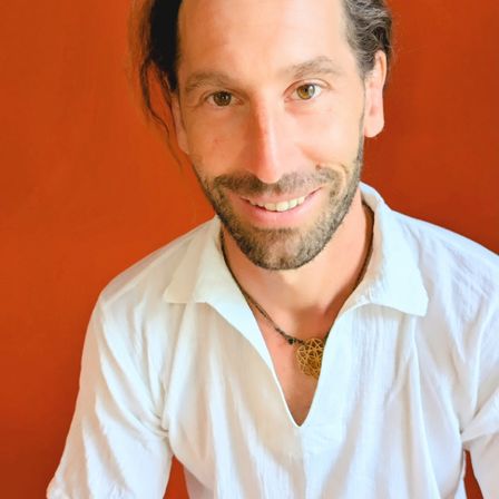 Ein lächelnder Mann mit Bart und Oberlippenbart, der ein weißes Hemd und eine goldene Anhängerkette trägt, posiert vor einem leuchtend orangefarbenen Hintergrund.