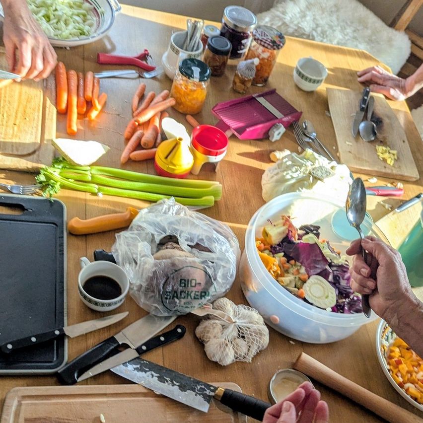 Ein Holztisch ist mit verschiedenen Gemüsesorten, Utensilien und Gläsern gefüllt. Es gibt Karotten, Sellerie und andere Gemüsesorten, zusammen mit Schüsseln, einer Reibe und einer Plastiktüte. Die Hände von Menschen sind ebenfalls sichtbar, die ein Messer und einen Löffel halten.
