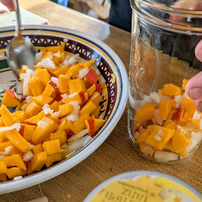 Eine Schüssel mit gewürfeltem Kürbis und Zwiebeln steht auf einem Holztisch neben einem Glas, das auch mit gewürfeltem Kürbis und Zwiebeln gefüllt ist. Eine Person hält einen Löffel und fügt möglicherweise weitere Zutaten hinzu.