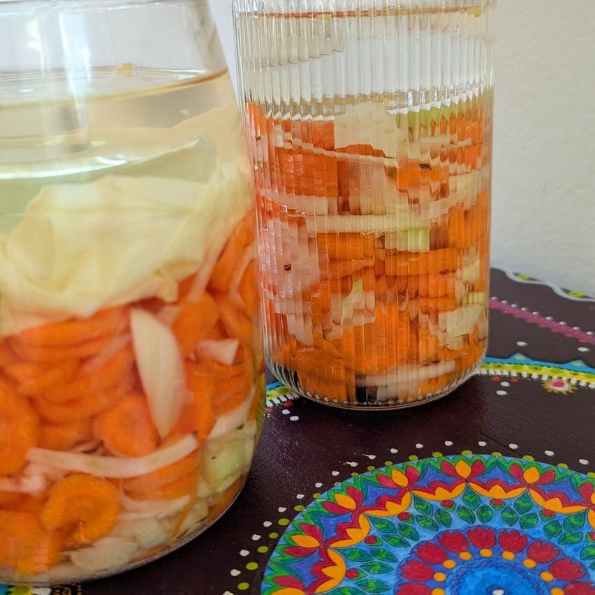 Zwei Glasgefäße gefüllt mit einer Mischung aus geschnittenen Karotten, Zwiebeln und Kraut, die in einer Flüssigkeit auf einem Tisch mit einem farbenfrohen Muster eingetaucht sind.