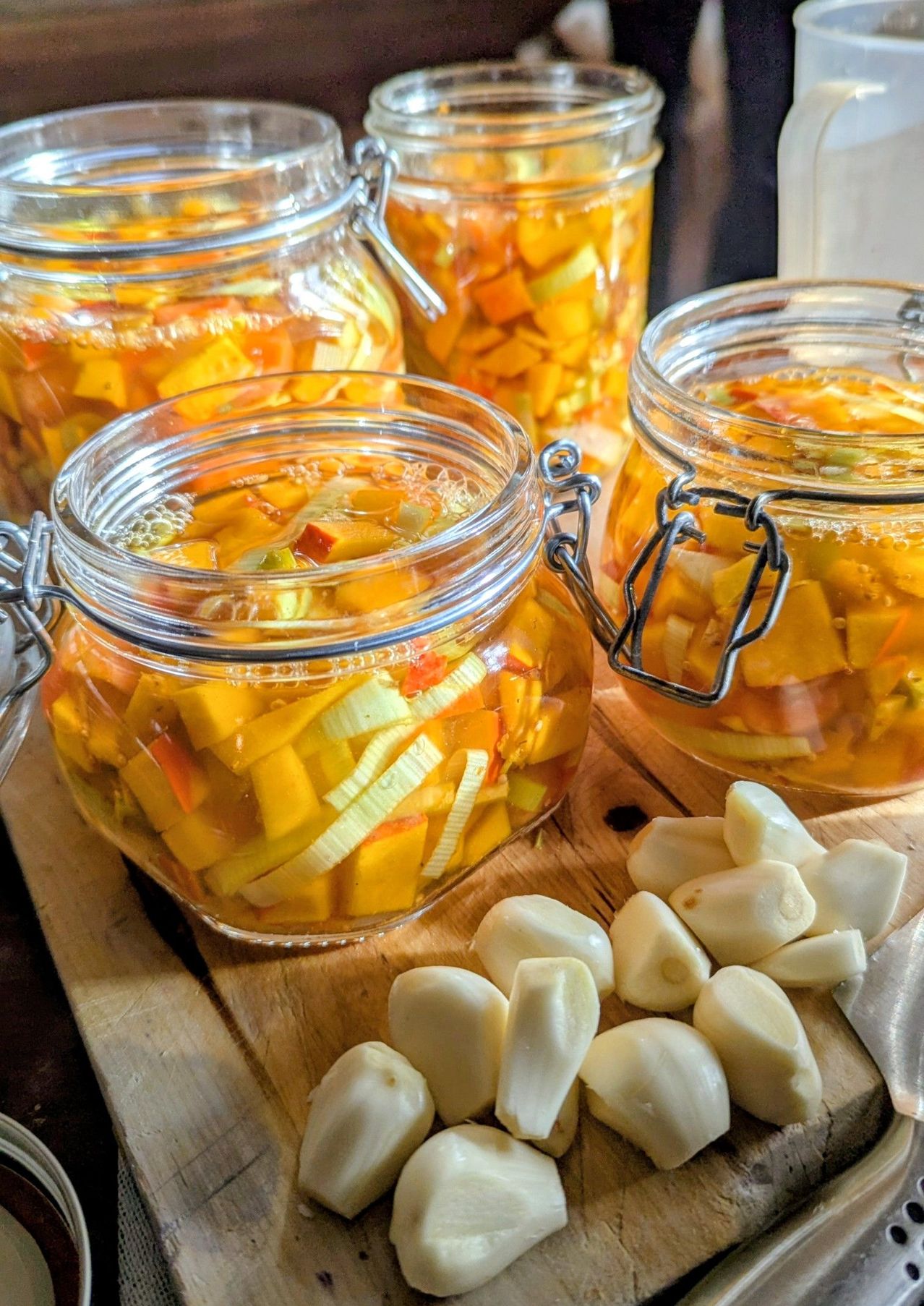 Mehrere Glasgläser mit eingelegtem Gemüse stehen auf einem Holzschneidebrett. Neben den Gläsern befinden sich mehrere Knoblauchzehen.