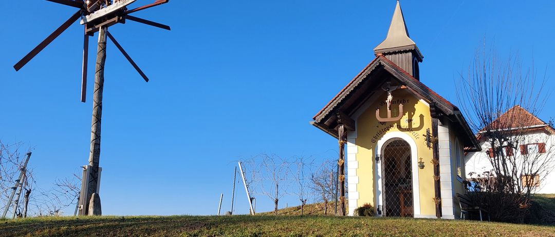 Eine Kapelle mit einem Turm und einem Kreuz steht auf einem Hügel. Ein Windrad mit roten Blättern steht davor. Die Kapelle ist gelb und weiß, und der Hügel ist grün.