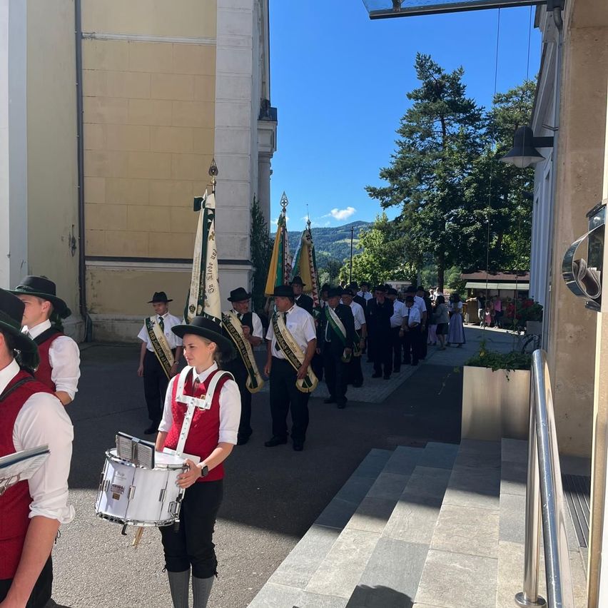 A marching band in traditional attire stands outdoors, with flags and a drummer leading the line. Behind them, a group of people watch from a building's side. Trees and mountains are visible in the background.