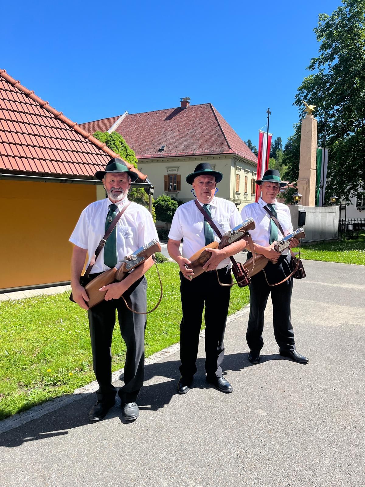 Three men, all wearing hats and uniforms, are standing on a street, holding rifles. A monument and a building with a flag are behind them.