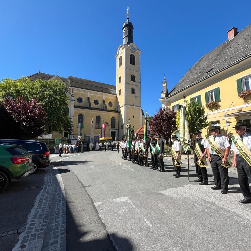 A group of people in uniforms are lined up outside a church. The street is lined with trees and parked cars.