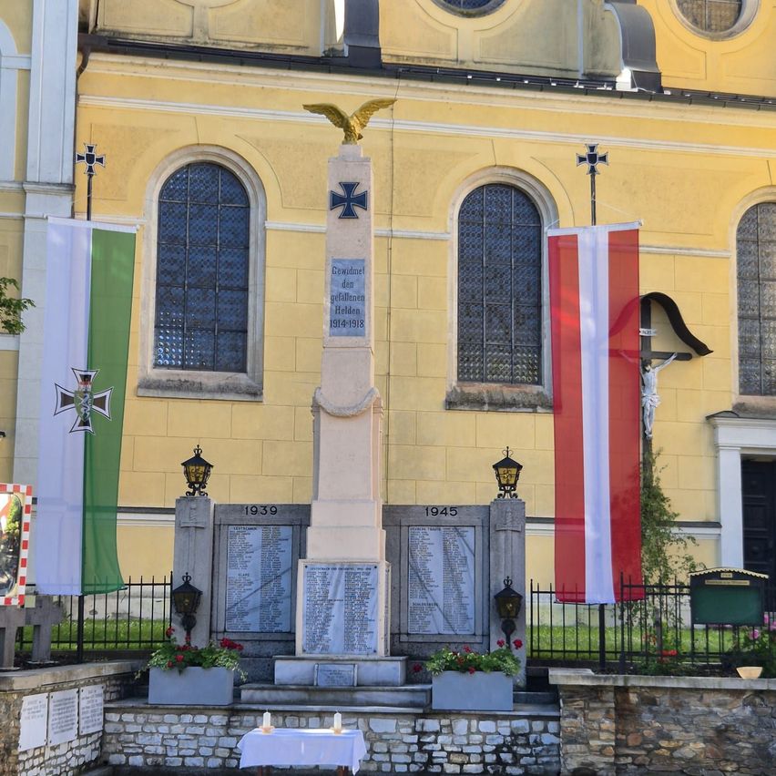 A memorial stands before a church, featuring an eagle and plaques with names from 1939 and 1945. Flags and lamps adorn the scene.