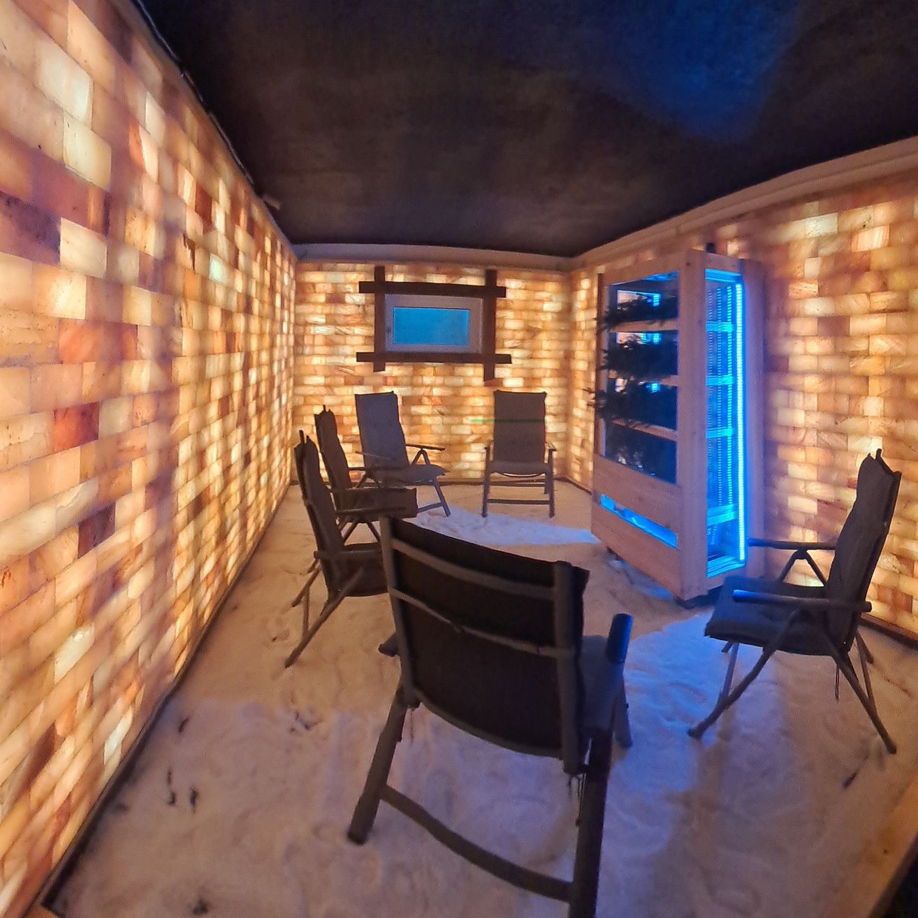 Ein kleiner Raum mit mehreren Stühlen, beleuchtet von blauem Licht, mit Wänden aus Salzziegeln und einem Kühlschrank.