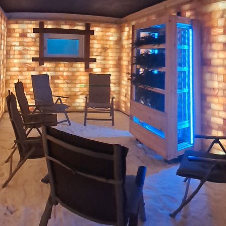 Ein kleiner, gemütlicher Raum mit steinernen Wänden, die von blauem Licht beleuchtet werden. Mehrere Stühle sind um einen kleinen Tisch herum angeordnet. Ein Kühlschrank mit blauer Beleuchtung steht rechts.