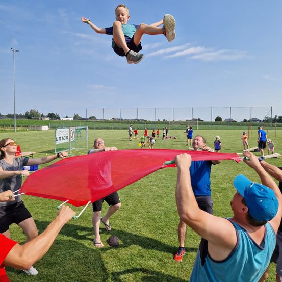 Ein Junge springt über ein rotes Netz, das von Erwachsenen auf einem Grasfeld gehalten wird, mit mehreren Menschen, die herumlaufen, und einer Umzäunung im Hintergrund.