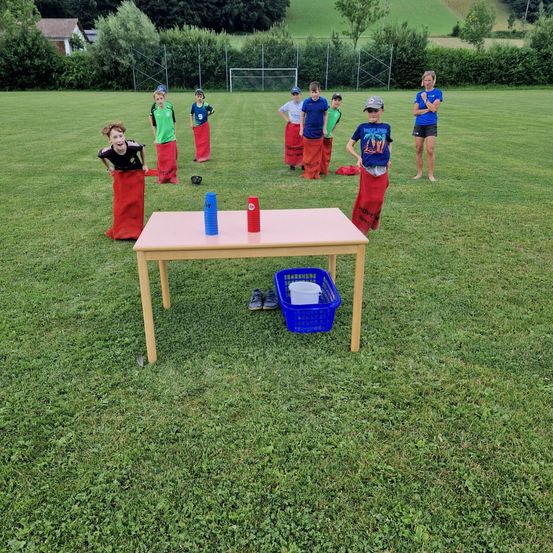 Mehrere Kinder, die Säcke über den Beinen tragen, sind auf einem Feld in einer Reihe aufgestellt. Eine Frau steht vor einem Tisch mit Tassen und einem Korb.