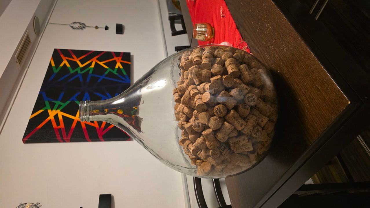 Eine große Glasflasche, gefüllt mit Weinkorken, steht auf einem Holztisch, mit einem farbenfrohen Gemälde im Hintergrund.