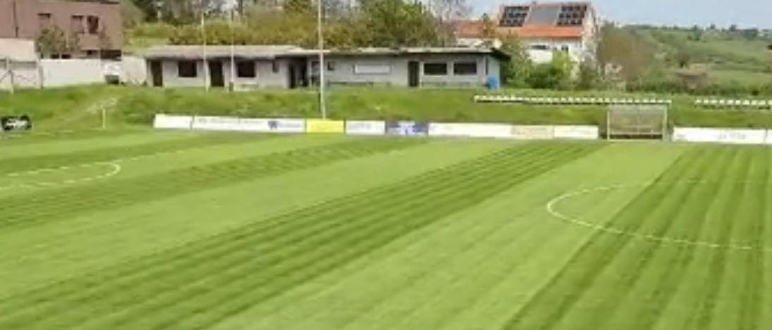 Eine breite Vogelperspektive auf ein grünes Fußballfeld mit einer Glasbarriere im Vordergrund, zwei Torpfosten am Ende und mehreren Gebäuden im Hintergrund.
