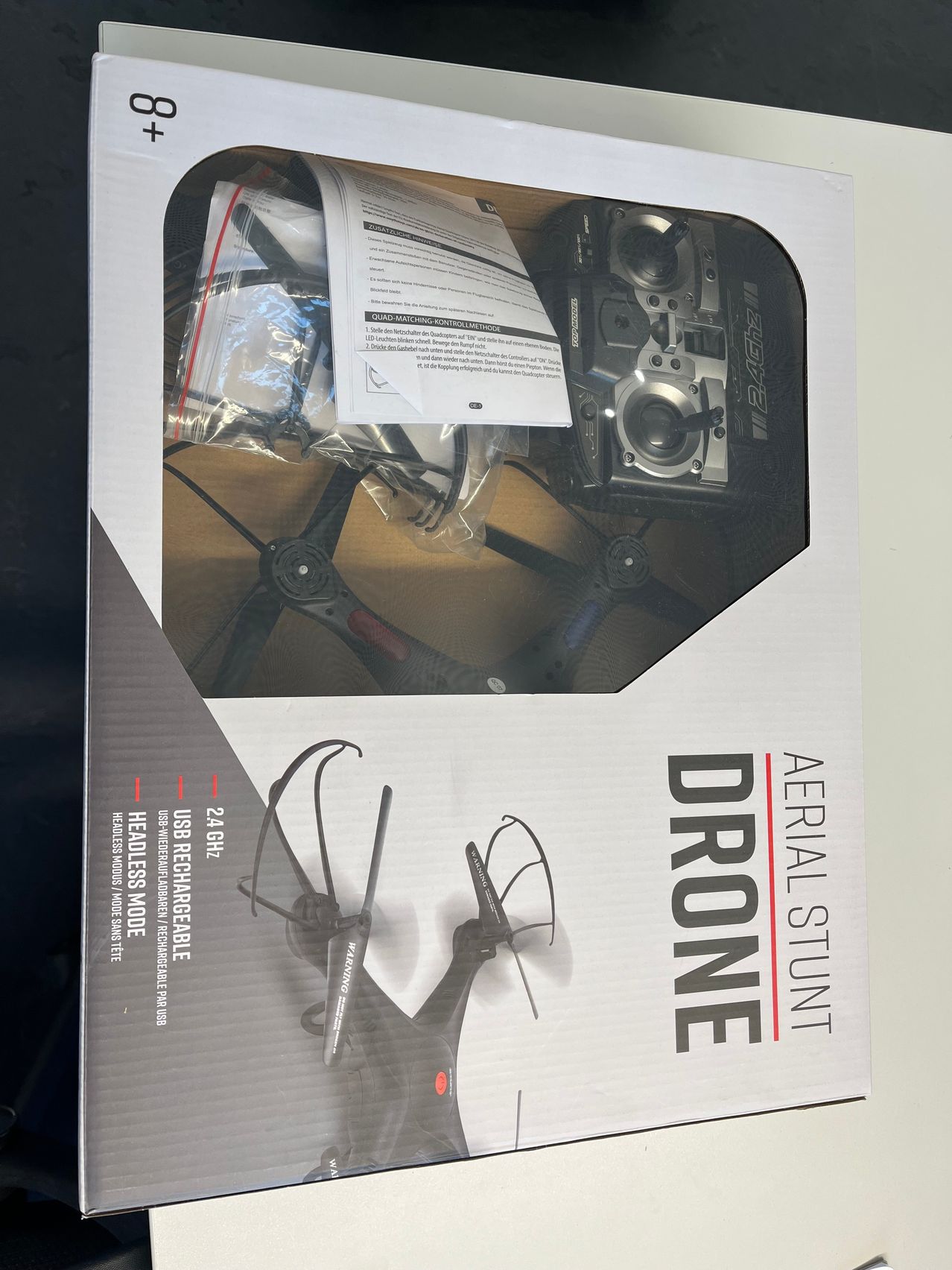 Die geöffnete Box eines Luftakrobaten-Drohnen mit sichtbarem Inhalt. Der Inhalt umfasst eine Fernbedienung, Propeller und eine Anleitung. Die Drohne ist auf der Box abgebildet zusammen mit ihren Funktionen.