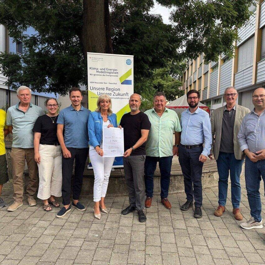 Eine Gruppe von Menschen steht vor einem Banner mit den Worten 'Unsere Region Unsere Zukunft'. Eine Frau in der Mitte hält ein Zertifikat. Hinter ihnen sind Gebäude und Bäume zu sehen.