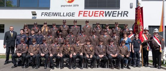 A group of volunteers in uniform poses for a photo in front of a building with a logo and the words 'Freiwillige Feuerwehr'.