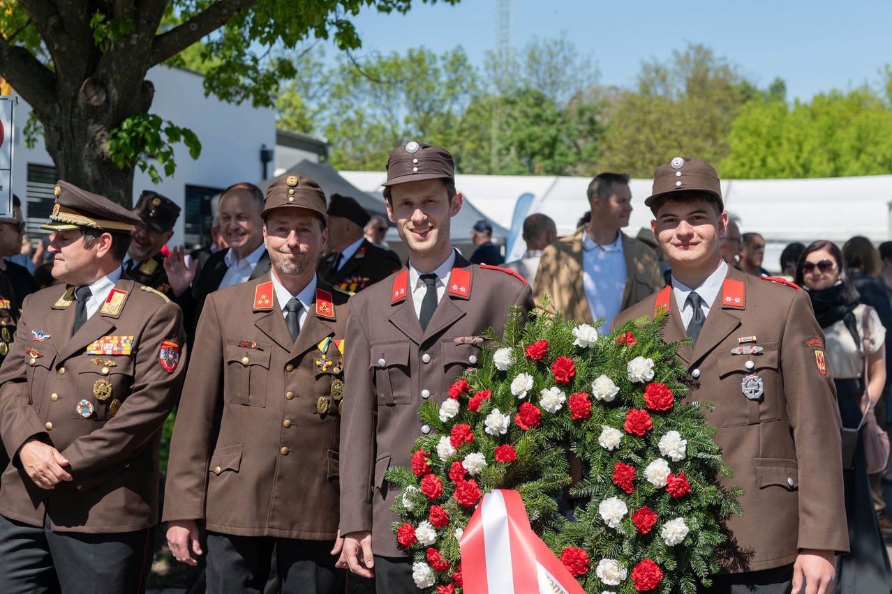 Four military men in uniforms stand in a row with a wreath in front of them. The wreath is red and white, with a red ribbon. Behind them, a crowd of people stand, some wearing hats. Trees and a white building are in the background.