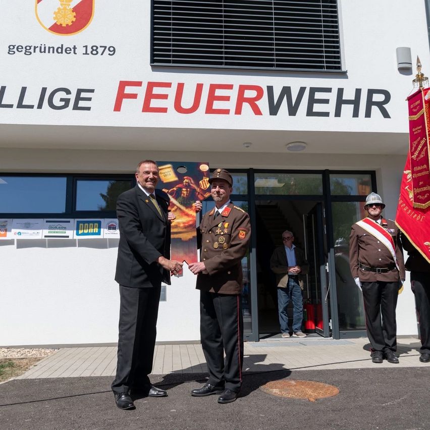 Two men are shaking hands in front of a building with a red flag and sign reading 'Feuerwehr'. They are holding a poster.