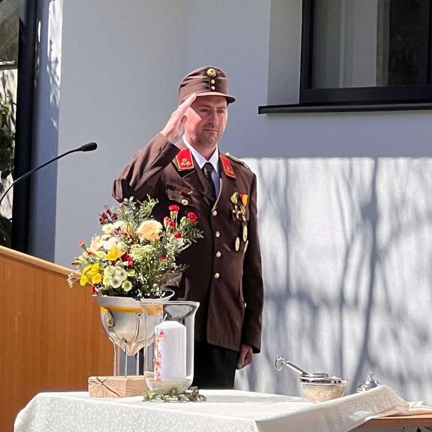 A man in a military uniform salutes while standing in front of a podium with a microphone. A flower arrangement and a candle are on the table in front of him.