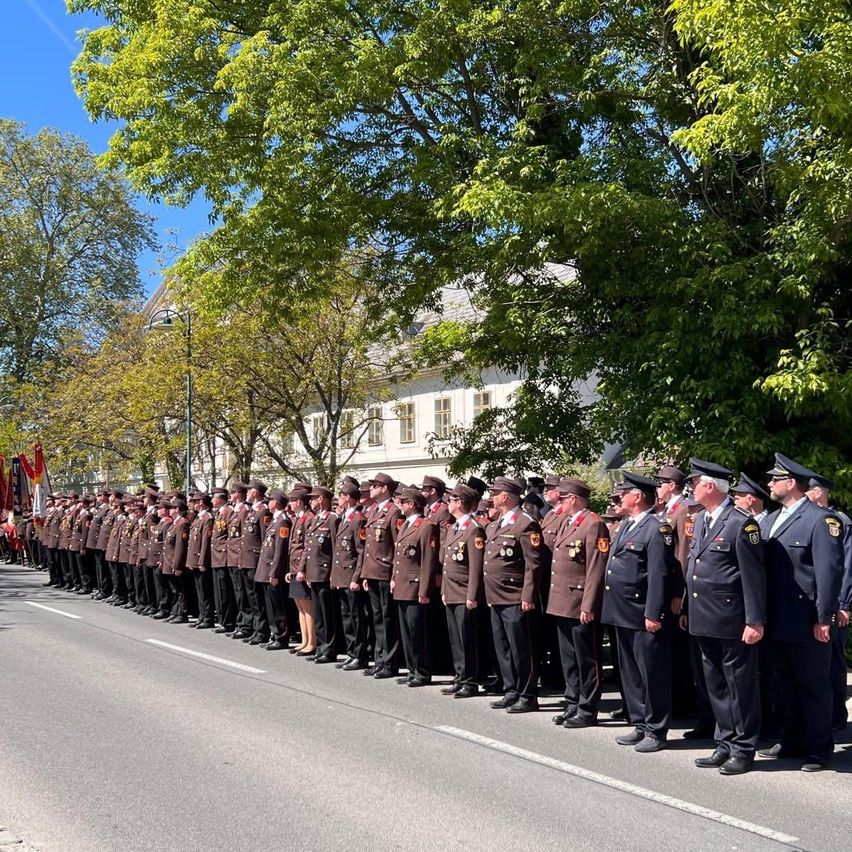 A line of uniformed individuals stands on the street, likely participating in a parade or ceremonial event, with trees and a white building in the background.