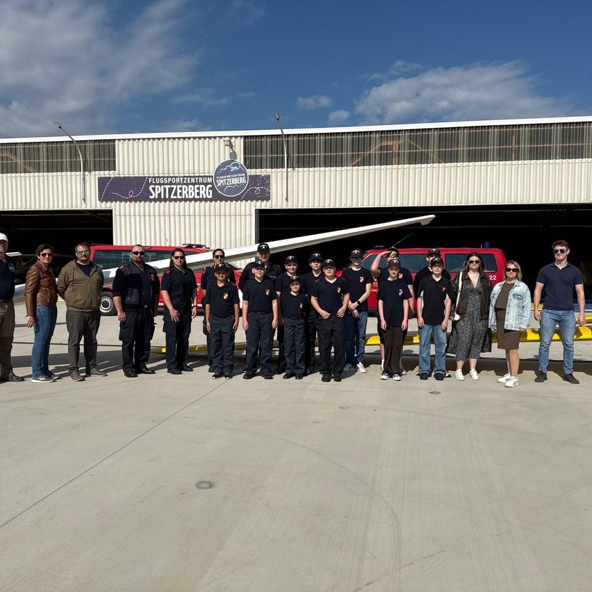 A group of people stand in front of a hangar, wearing uniforms, with a plane parked in the foreground.