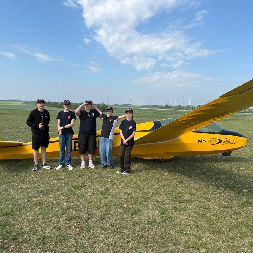 Five individuals stand in front of a yellow glider, smiling and posing for a photo. They wear casual attire, and the glider is marked with black and yellow. Behind them is a grassy field with distant windmills.