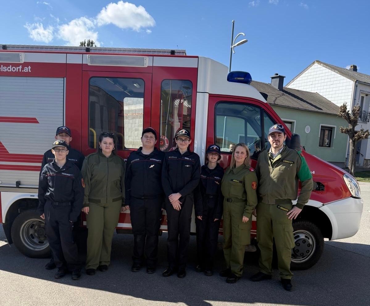 Eine Gruppe uniformierter Personen, wahrscheinlich Feuerwehrleute, posiert für ein Foto vor einem roten und weißen Feuerwehrwagen. Sie stehen vor einem Haus mit Schornstein.