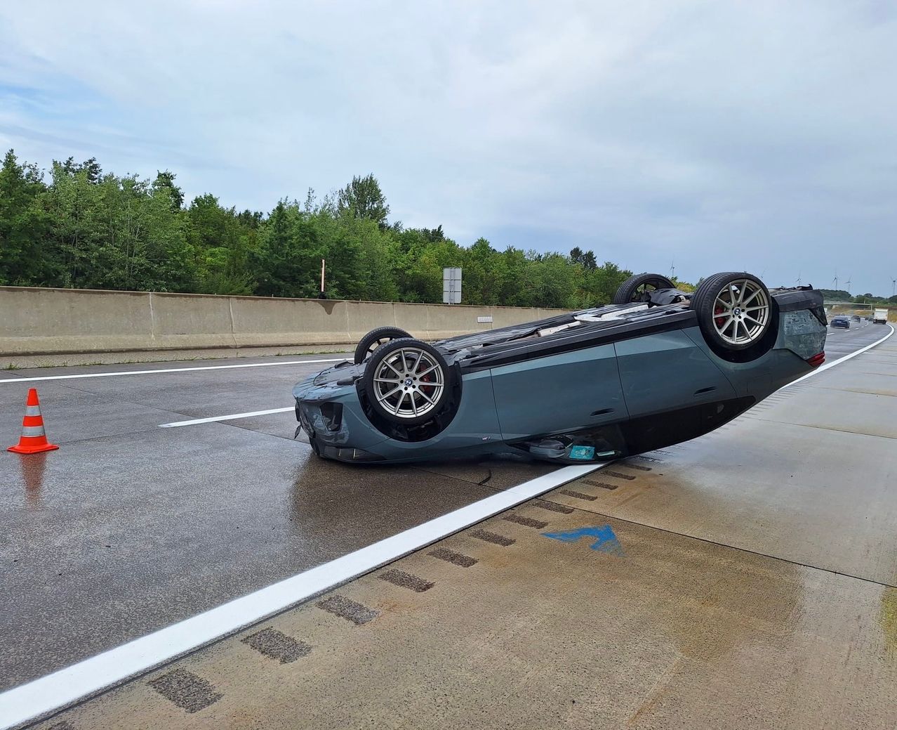 A car is flipped on its side on a wet road, with tires in the air and a blue arrow pointing left.