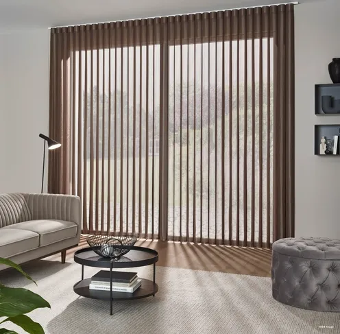 Ein Wohnzimmer mit einer beigen Couch, einem Couchtisch und einer modernen Lampe. Der Raum verfügt über große vertikale Jalousien, einen beigen Teppich und Regale mit Dekorationsartikeln. Draußen ist ein Blick auf Bäume zu sehen.