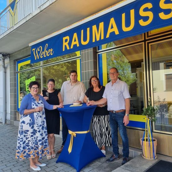 Fünf Personen stehen vor einem Geschäft namens Weber Raumauss, halten Gläser und eine Torte. Das Geschäft hat ein blaues Banner, Glasfenster und einen Topf mit einer Pflanze.