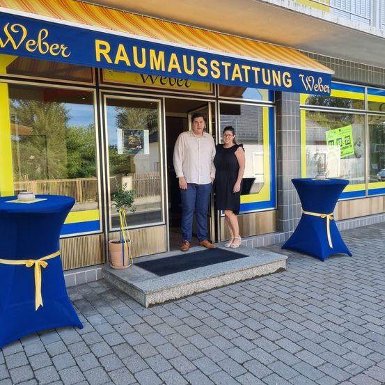 Zwei Personen stehen vor einem Geschäft mit einem blau-gelben Vordach. Der Mann trägt ein weißes Hemd und blaue Hose, die Frau ein schwarzes Kleid. Sie lächeln beide.