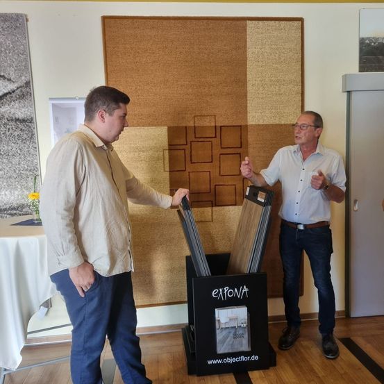 Zwei Männer diskutieren Produkte in einem Raum mit einer großen braun gemusterten Wand, einem Tisch mit einer Blumenvase und einem Holzboden.