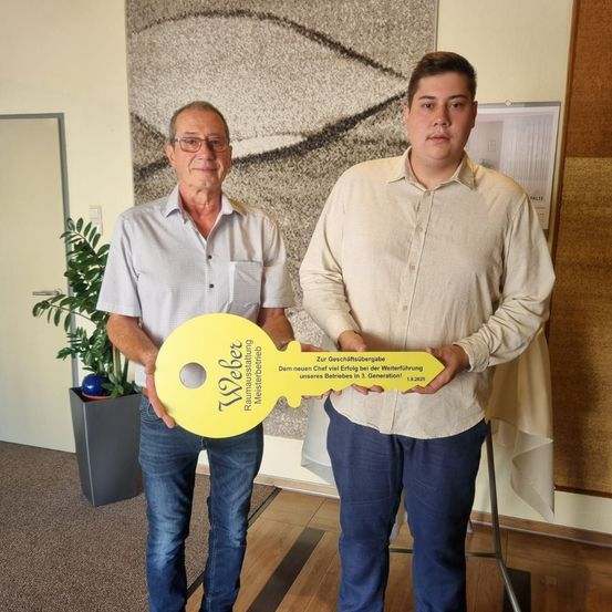 Zwei Männer halten einen großen gelben Schlüssel, einer trägt eine Brille und ein graues Hemd, der andere trägt ein helles Hemd und Jeans. Sie stehen in einem Raum mit Wandbild, Pflanze und Kalender.