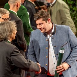 Ein Mann in einem blauen Jackett schüttelt die Hand eines älteren Mannes in einem grauen Anzug. Sie lächeln beide, und der Mann im blauen Jackett hält eine gerollte grüne Fahne. Dahinter stehen mehrere Leute und beobachten.