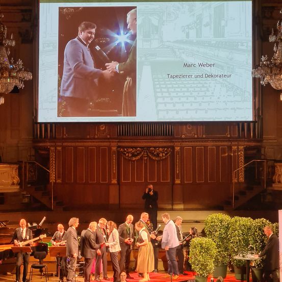 Eine Preisverleihung findet in einem großen Saal mit Bühne, Leinwand und Kronleuchtern statt. Die Leinwand zeigt Marc Weber, einen Tapezierer und Dekorateur, der jemandem die Hand schüttelt. Eine Person auf der Bühne macht Fotos.