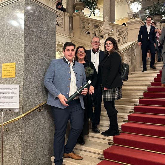 Eine Gruppe von Menschen steht auf einer Treppe, möglicherweise in einem historischen Gebäude. Ein Mann hält eine Schriftrolle, und zwei Frauen stehen neben ihm. Hinter ihnen läuft ein Mann die Treppe hoch.