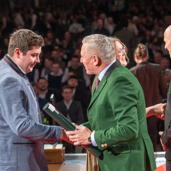 Ein Mann in einem grünen Anzug überreicht einem jungen Mann in einem blauen Anzug einen Preis, vor Publikum im Hintergrund.