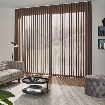 Ein Wohnzimmer mit vertikalen Jalousien, einem beigen Sofa, einem runden Tisch mit Büchern und einer Lampe. Eine schwarze Vase und Bücher stehen auf den Wandregalen.