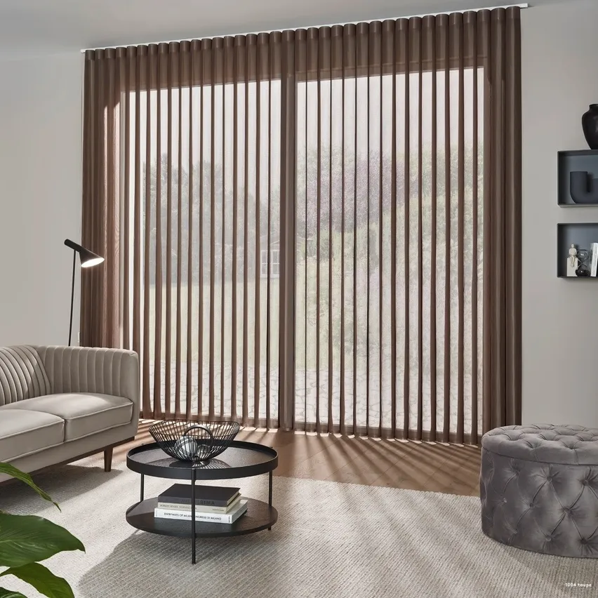 Ein Wohnzimmer mit vertikalen Jalousien, einem beigen Sofa, einem runden Tisch mit Büchern und einer Lampe. Eine schwarze Vase und Bücher stehen auf den Wandregalen.