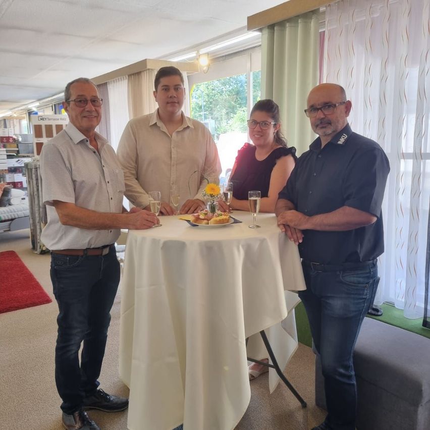 Vier Personen stehen um einen Tisch mit Weingläsern und einem Teller mit Essen. Sie lächeln und scheinen zu feiern.