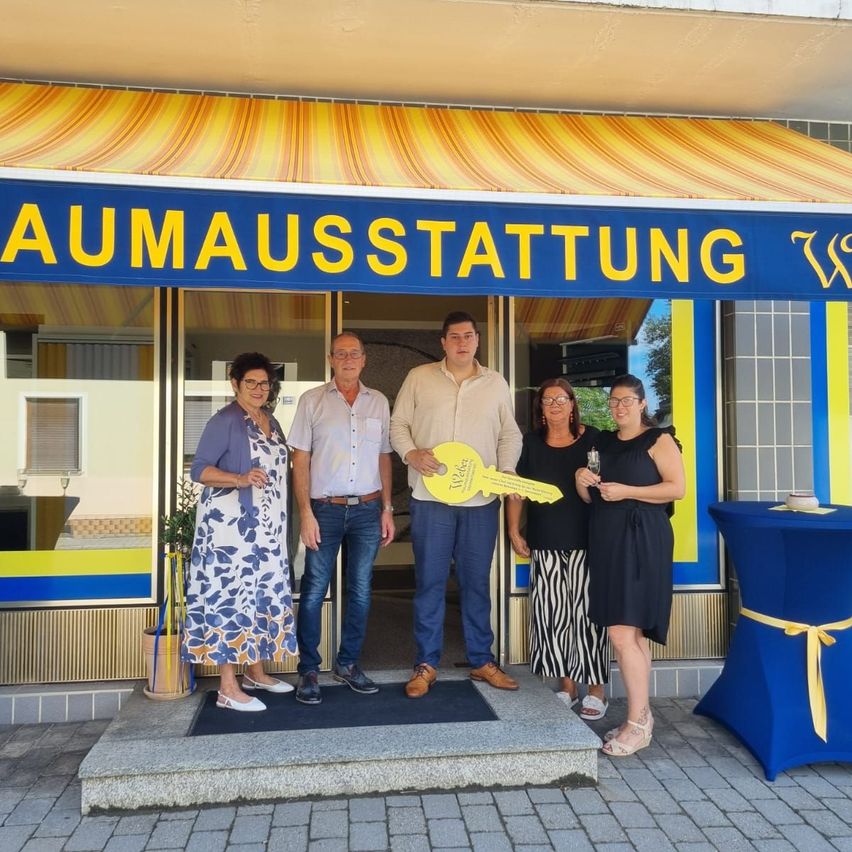 Fünf Personen stehen vor einem Geschäft namens Baumausstatting. Sie halten einen Schlüssel und Champagner in den Händen. Sie lächeln und posieren für ein Foto.
