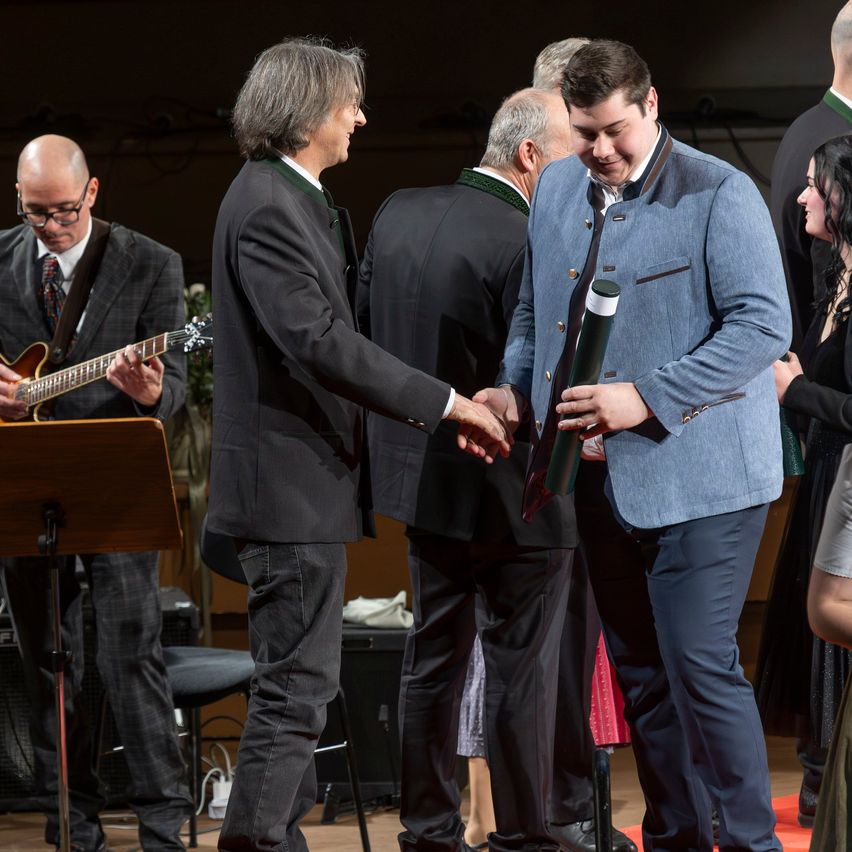 Ein Mann erhält von einem anderen Mann eine Auszeichnung, während er auf einer Bühne vor einer Gruppe von Menschen steht. Ein Gitarrist spielt auf der linken Seite der Bühne.