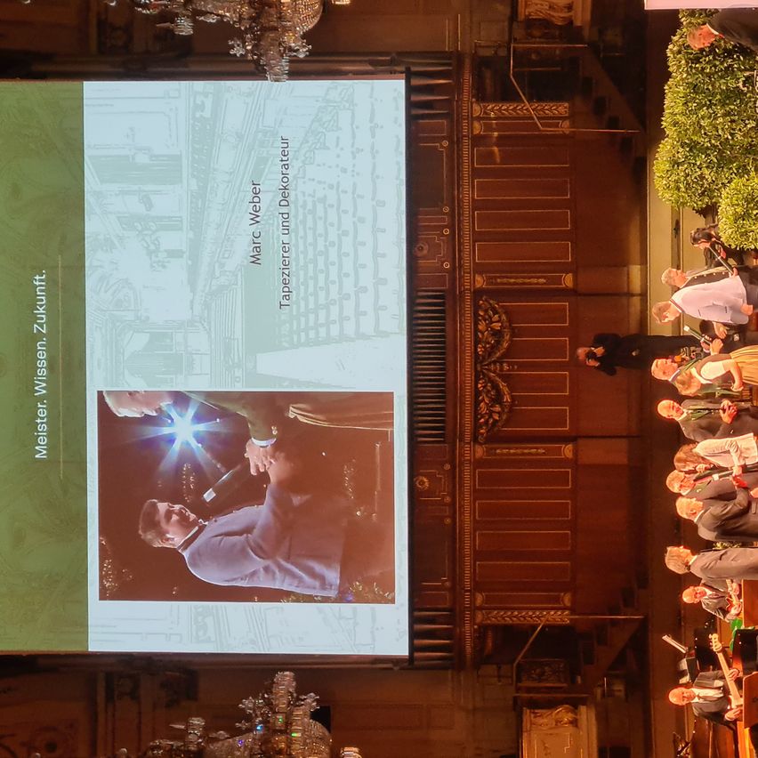 Eine große Leinwand zeigt ein Bild von Marc Weber, einem Tapezierer und Dekorateur, mit einer Person, die ein Mikrofon hält. Mehrere Personen sitzen und stehen hinter der Leinwand.
