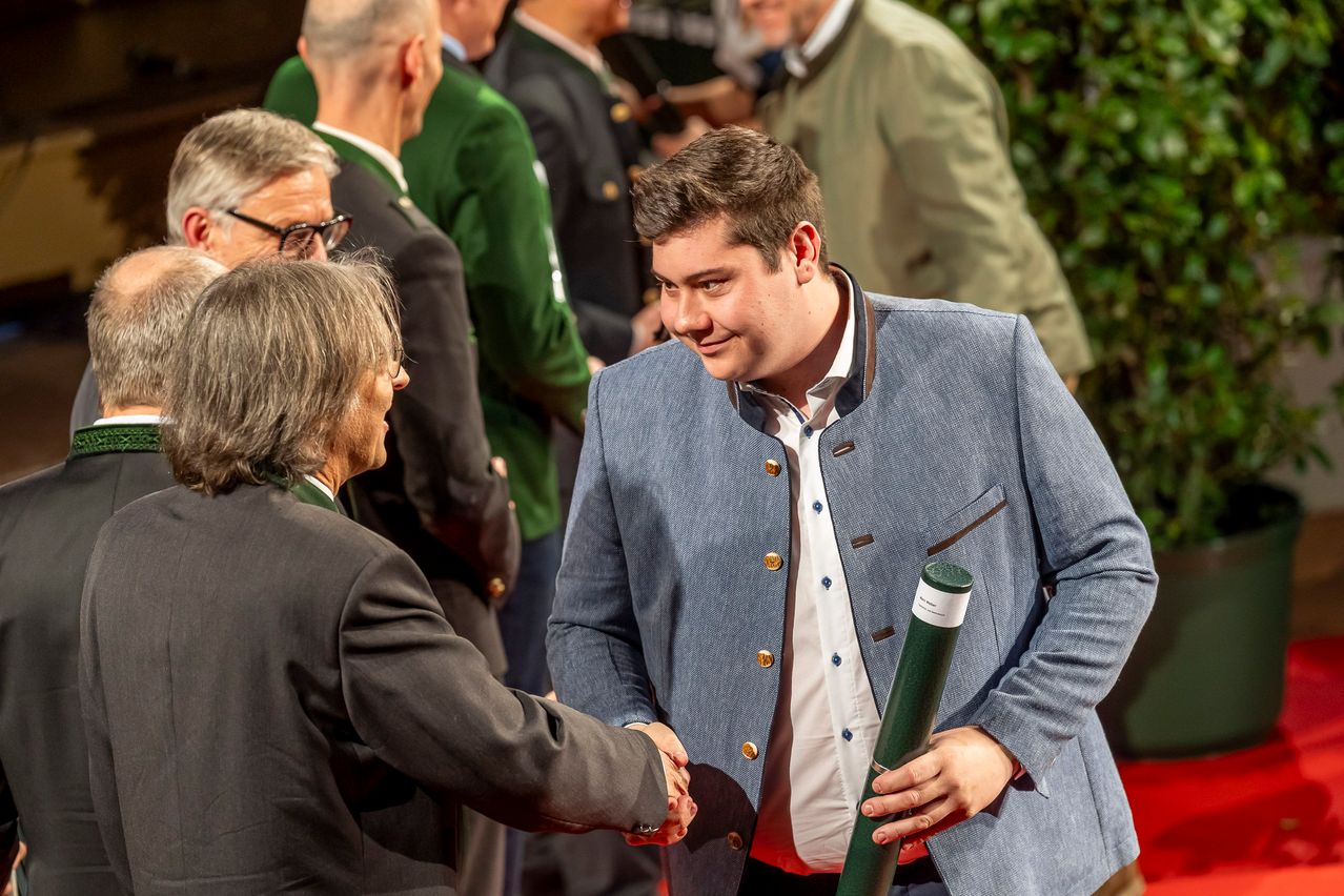 Ein junger Mann in einem blauen Mantel hält ein Diplom in der Hand und schüttelt einem älteren Mann in einem grauen Anzug die Hand. Dahinter steht eine Menschenmenge auf einem roten Teppich, einige tragen grüne Jacken.