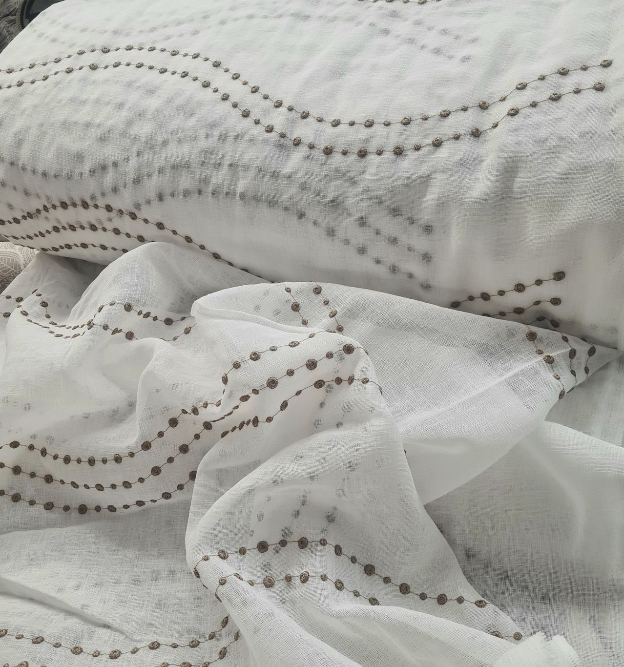 Eine Nahaufnahme eines weißen Stoffs mit einem welligen braunen Perlenmuster auf grauem Hintergrund. Der Stoff scheint eine Bettwäsche oder ein Kissenbezug zu sein.