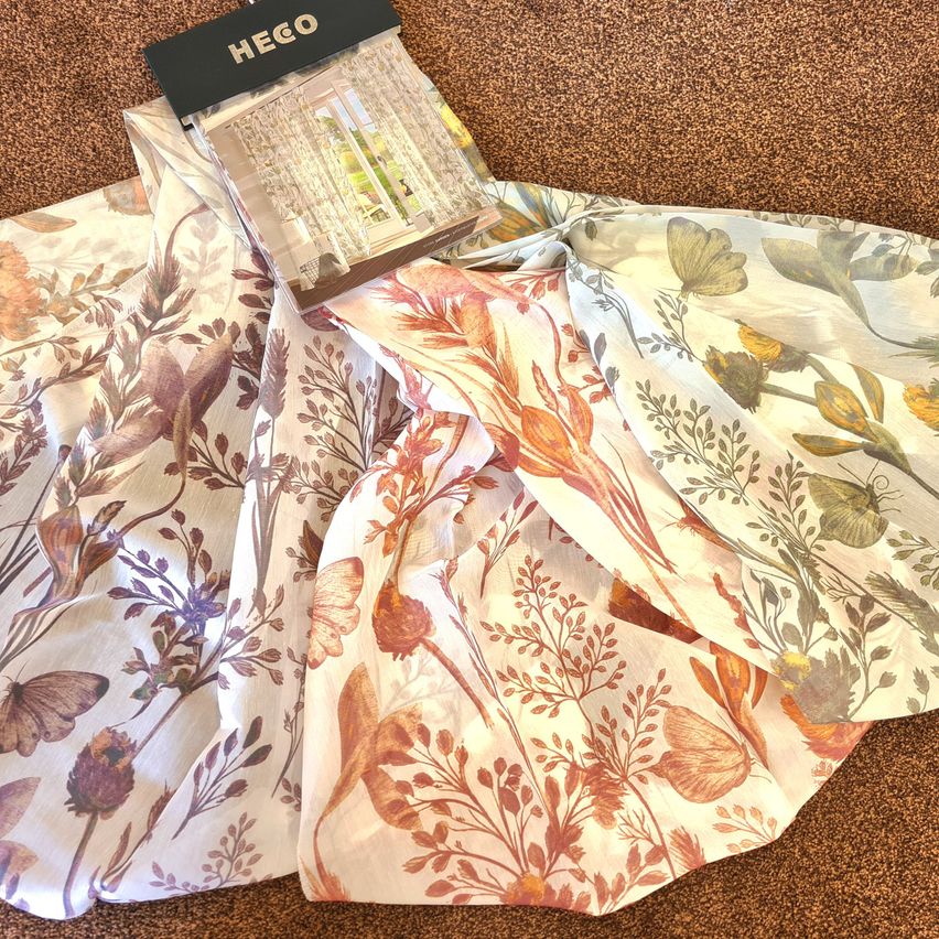 Ein floraler Vorhang mit Schmetterlingsdesign ist auf einem braunen Teppich gefaltet. Der Vorhang ist weiß mit orangenen, lila und grünen Blumen. Ein schwarzer HEGO-Anhänger befindet sich am Vorhang.