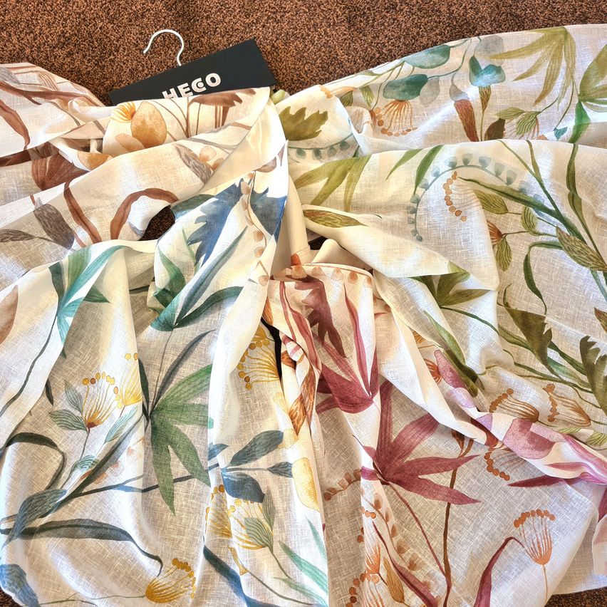Ein floral gemusterter Stoff mit verschiedenen Blättern und Blumen hängt an einem Kleiderbügel. Das Design ist auf einem weißen, leicht strukturierten Material, möglicherweise Leinen, gedruckt.